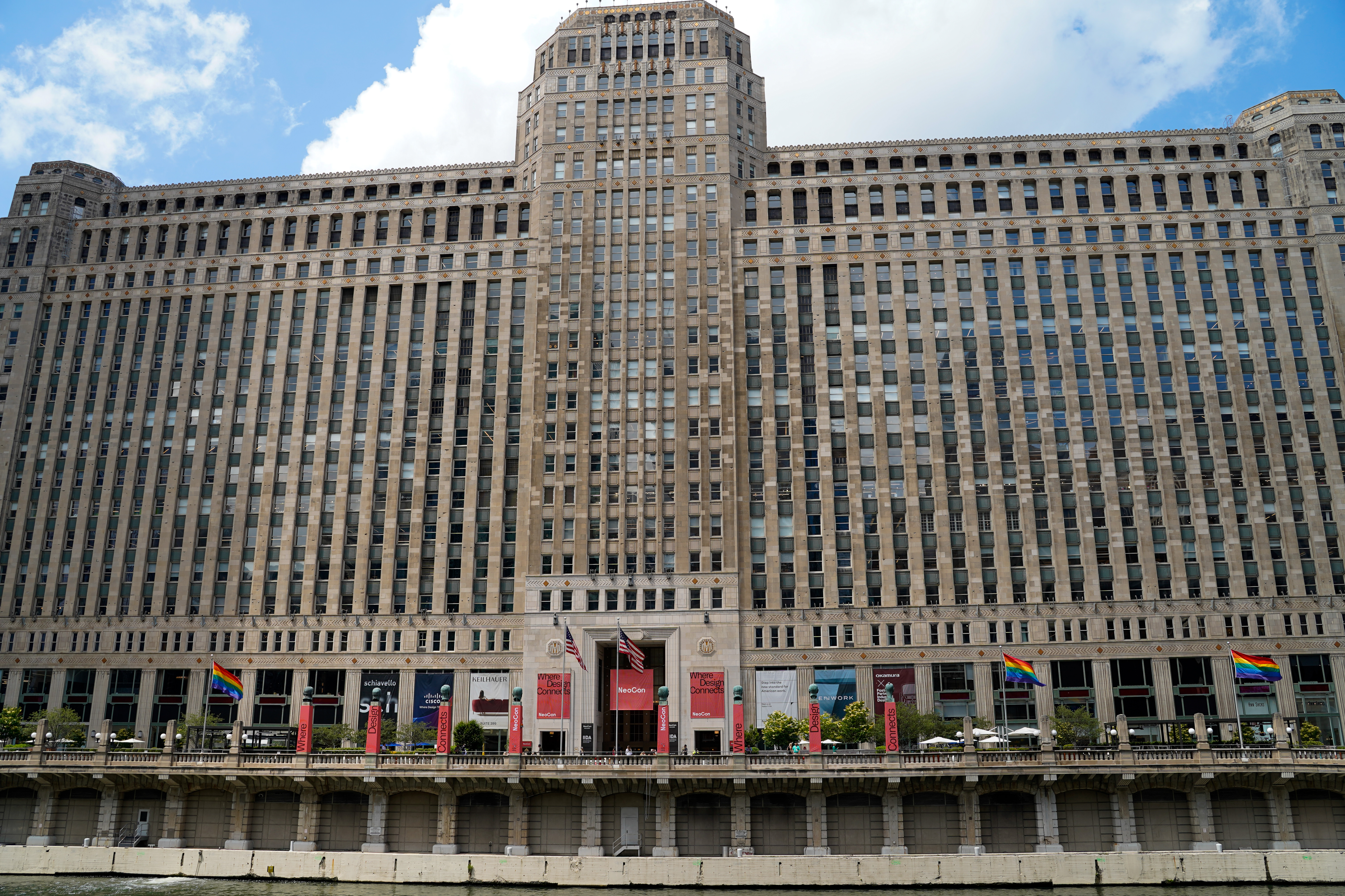 Historic Merchandise Mart building in Chicago viewed from across the Chicago River during NeoCon 2026. The massive Art Deco facade fills the frame, with NeoCon banners, American flags, and rainbow flags displayed along the riverfront beneath a bright blue sky with scattered clouds.