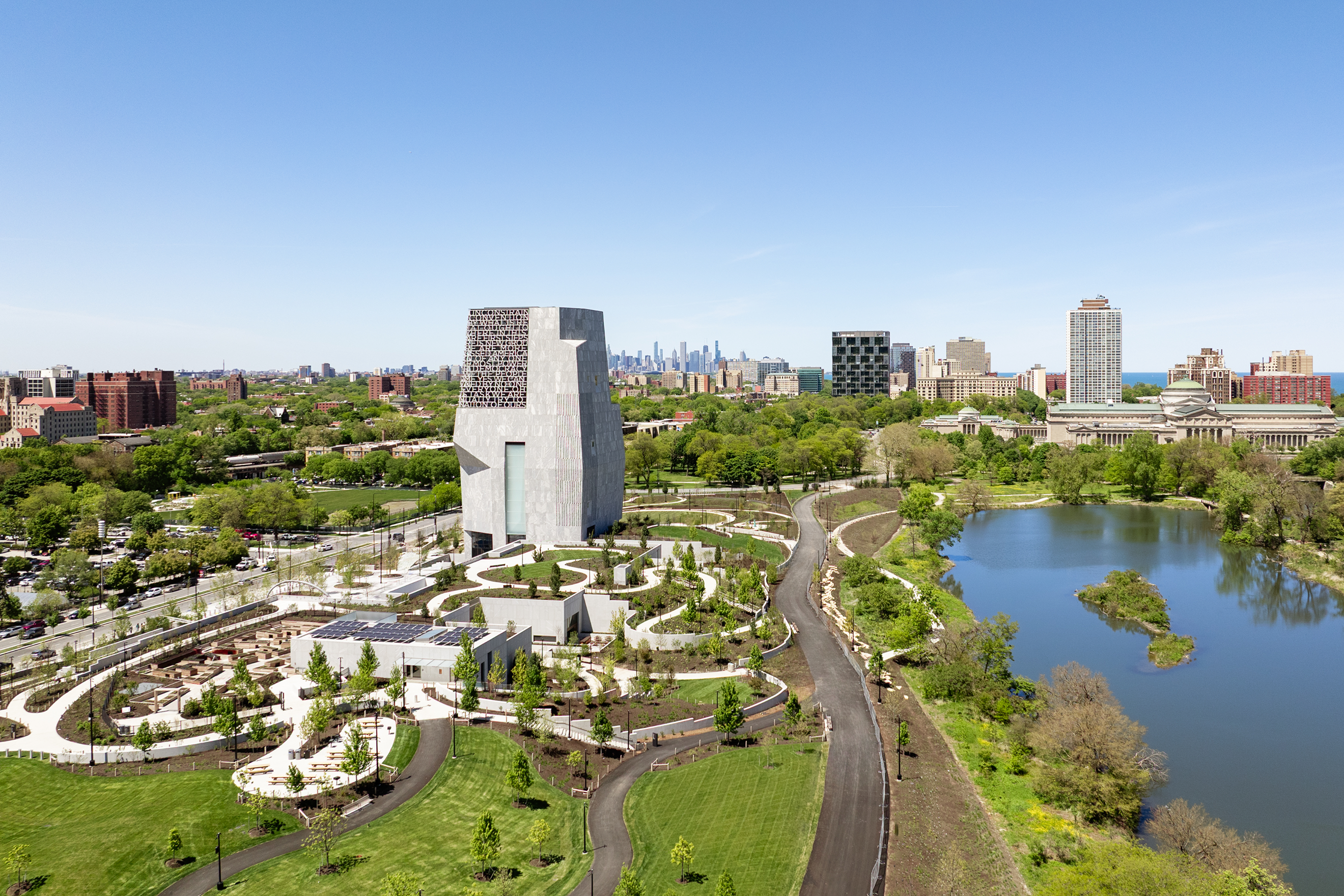 Aerial view of the Obama Presidential Center in Chicago surrounded by landscaped gardens, walking paths, and a large pond on a clear sunny day. The museum tower rises above Jackson Park with the Chicago skyline visible in the distance, highlighting the scale of the campus and its connection to the surrounding parkland.
