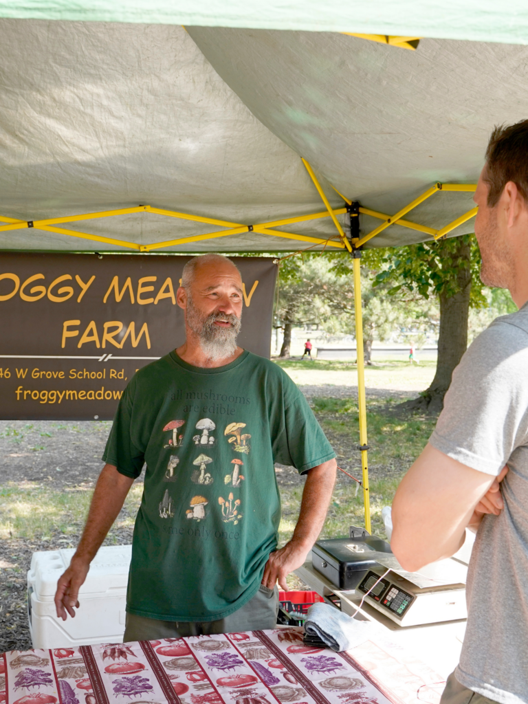 Farmer speaking with Paramount Evetns Chef at an outdoor market stand. A banner behind him reads "Froggy Meadow Farm."