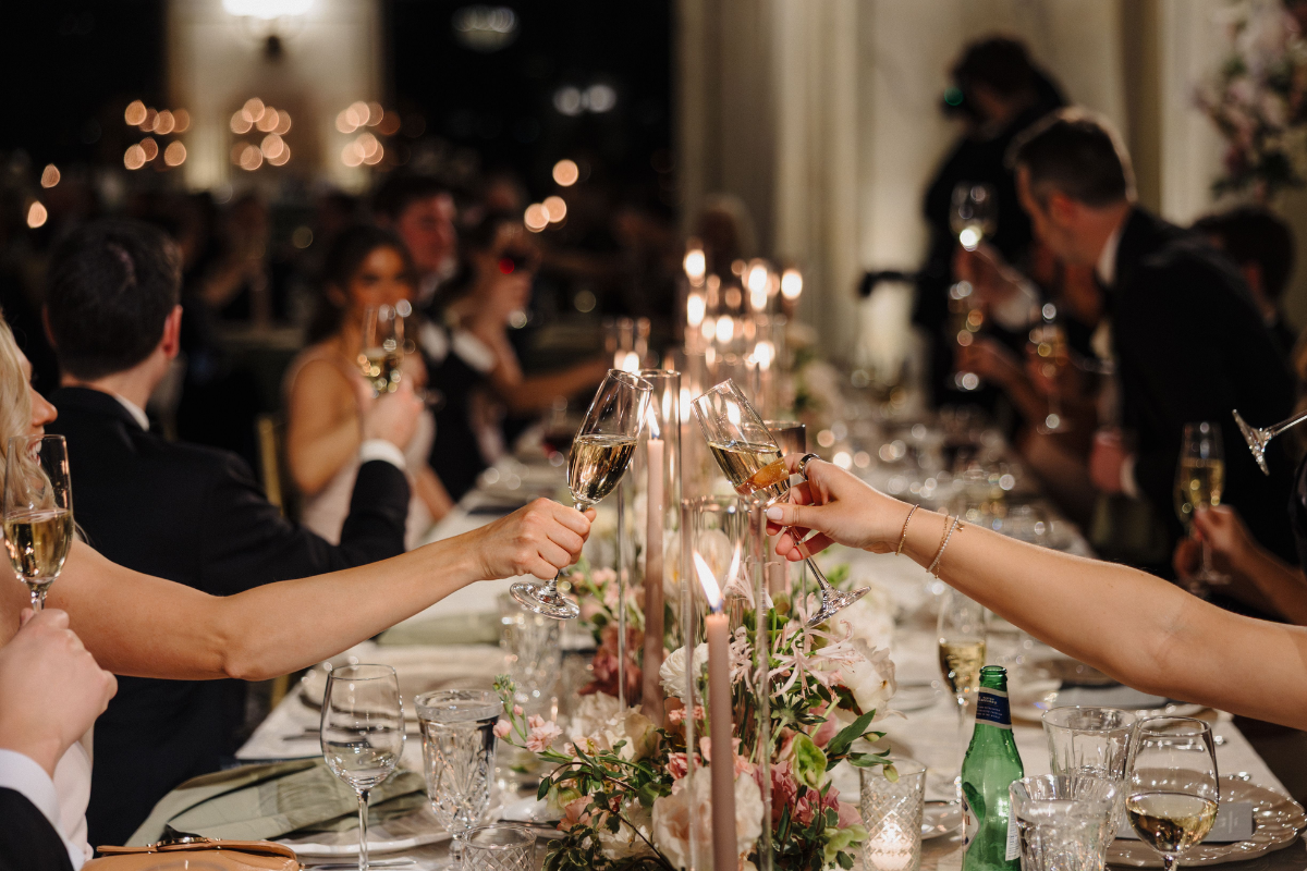 Wedding guests raise champagne glasses for a toast around a candlelit reception table decorated with lush floral centerpieces. The warm lighting captures the inviting atmosphere of a fall or winter wedding celebration.