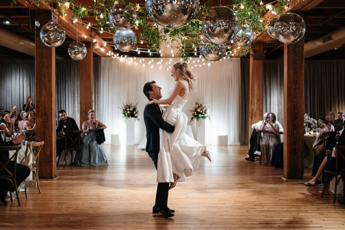 Newlyweds laugh as the groom lifts the bride during their reception beneath hanging disco balls greenery and string lights. Guests watch from both sides of the dance floor during the joyful 2027 Chicago wedding celebration.