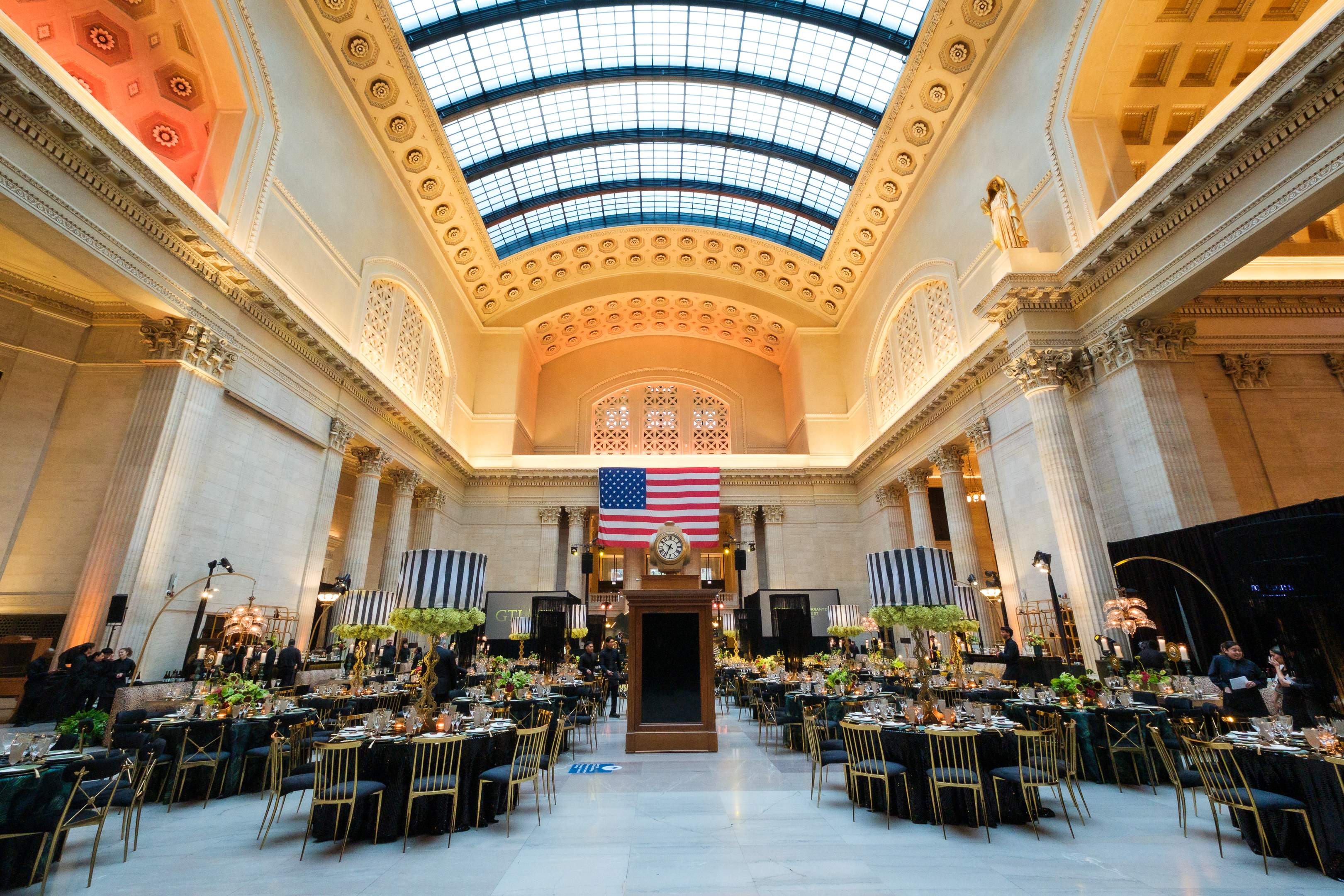 Grand historic event hall with towering marble columns, an arched glass ceiling, and an American flag above a central clock. Round tables with black linens, gold chairs, and elaborate floral centerpieces are arranged for a formal corporate event.